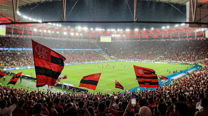 Vista em nível de campo do gramado do Maracanã à noite, com as luzes dos refletores e a torcida desfocada ao fundo. Um jogador do Flamengo em movimento e um do Cruzeiro em postura defensiva estão centralizados na ação. Imagem limpa, sem gráficos. Transmissão Flamengo x Cruzeiro GE TV YouTube ao vivo